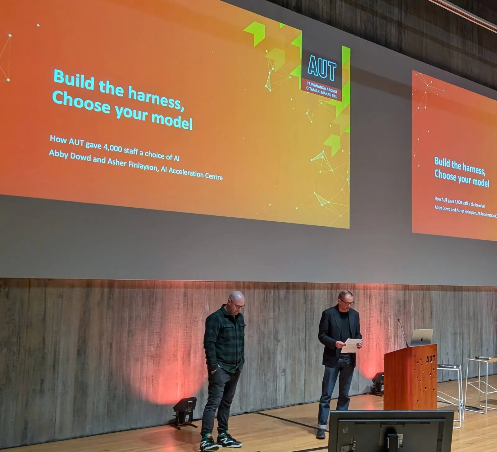 Abby Dowd and Asher Finlayson on stage under their title slide, “Build the harness, choose your model: how AUT gave 4,000 staff a choice of AI”